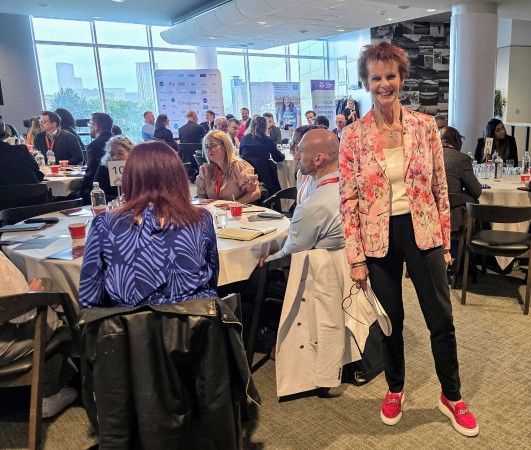 Rt. Hon. Anne Milton stands in a busy room of SRAG attendees at tables. She is smiling and projecting positivity that reflects the atmosphere on the day. 